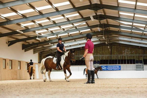 Equine lecturer supervises a riding lesson in indoor arena