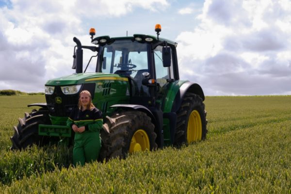 Girl in green overalls standing in front of a green tractor in a field.